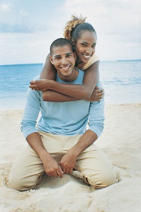 Young black couple on sandy beach. Both are smiling and kneeling on the sand. She has her arms wrapped around his shoulders from behind him.