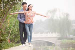 Young Asian couple standing on a garden wall near a river. She is in front of him with her arms spread as if to fly. He has his hands on her hips to balance her.