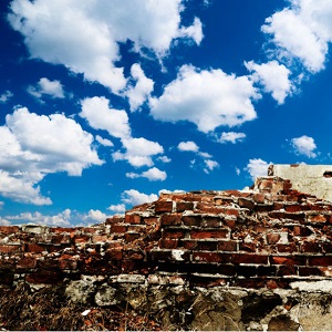 A crumbling brick wall is shown with a blue peaceful sky peeking over the top.
