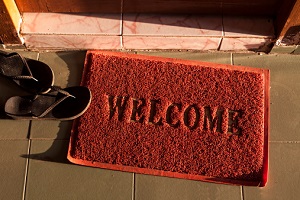 A red welcome doormat sits on a porch with a flip-flop laying on its edge.