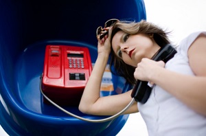 Young woman in white tee shirt leaning against phone booth, holding payphone in left hand. She has her hand on her forehead and looks upset and stressed.