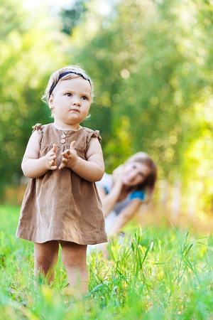 Toddler standing in grass with mother in background