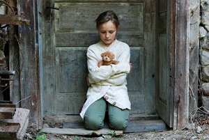 Child crouching in door of abandoned house with teddy bear