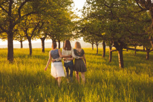 Rear view photo of three people with long hair walking in grassy space under trees arm in arm