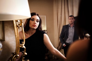 Unhappy looking woman wearing black dress looks in  mirror while a man stands in the background