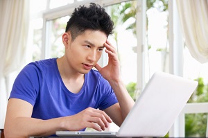 Worried Young Man Sitting At Desk Using Laptop At Home