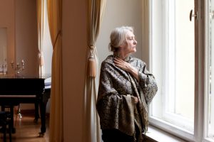 Senior adult wearing elegant brocade wrap with white hair in a clip stands at window looking outside