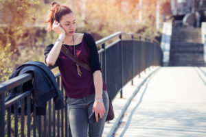 Young adult with ponytail leans on rail on street, looking down to the side, holding phone
