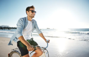 adult with facial hair, short hair, rides bike along shore smiling