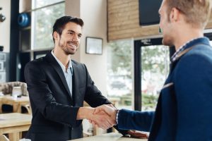 Two people in suits shake hands, smiling, in a cafe