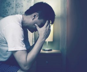 Muted tone photo of person sitting on edge of bed leans over with face in hands, upset