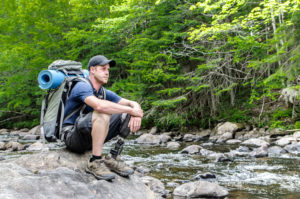 Person seen from side, with prosthetic leg and backpack sitting on rock by the river during a summer day