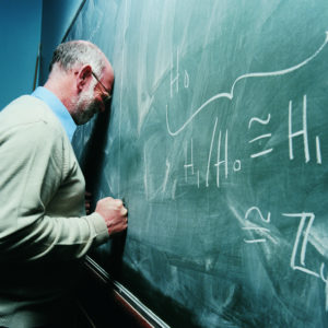 Person in classroom wearing shirt, cardigan, and tie presses head against blackboard