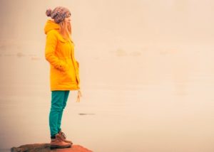 Person with long hair wearing had and down jacket stands on rock looking out to water