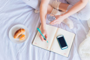 Cropped photo of crossed legs of person on bed with breakfast writing in scheduling book