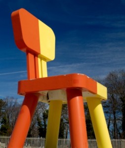 A huge chair towers over trees and a wooden fence, against a rich blue sky.