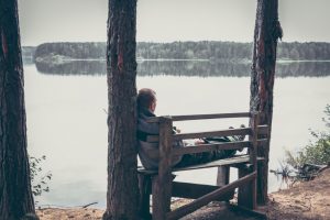 Pensive young person on the high edge of river bank sitting on bench and looking out at tranquil water