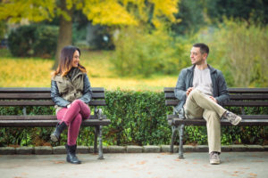 Two young people sit on benches in park looking at each other
