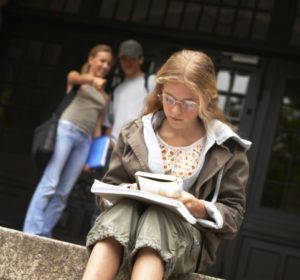 Blonde preteen sits on school steps reading while kids behind point