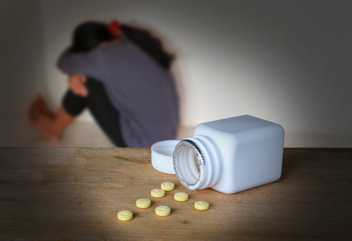 A spilled bottle of pills on a table in front of a woman in the corner