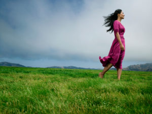 Person wearing long fuchsia dress stands barefoot in grassy field while storm clouds gather