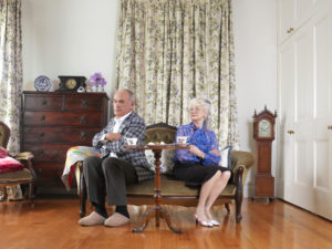 Two older adults sit on sofa at small table facing away from each other with arms crossed