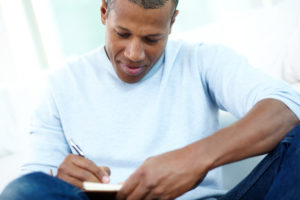 Person in casual clothes sits cross-legged and writes notes in notebook, thinking