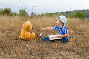 Child reads book with stuffed bear in field