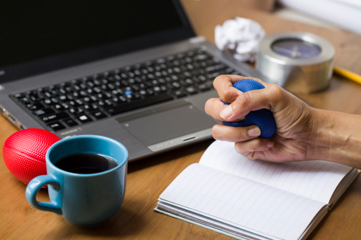 Hand squeezing stress ball on desk with computer and coffee