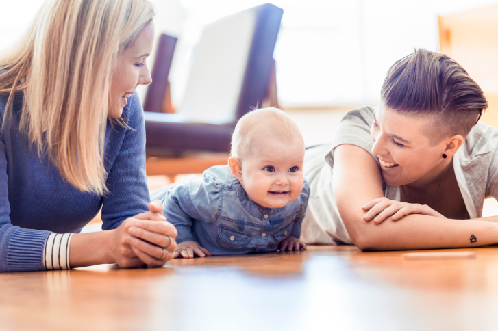 Two mothers smiling at baby on wood floor