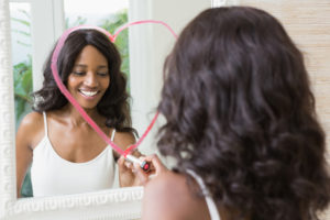 Young adult with curly long hair draws heart on mirror with lipstick, smiling