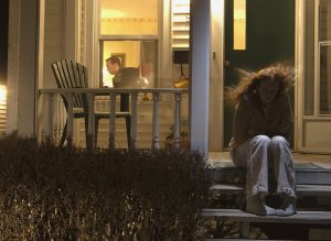 Man works on computer inside while sad woman sits on porch