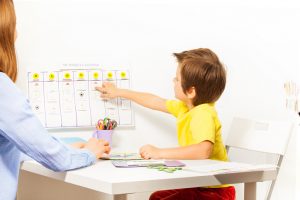 Young boy sits with parent at small table and adds activity to calendar on wall