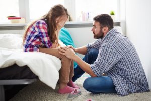 Father sitting on floor offers support to grieving teen sitting on bed
