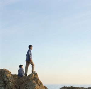 Man stands on rock cliff looking out to see while child sits behind him