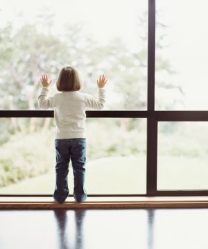 Standing child looks out of window, hands on glass