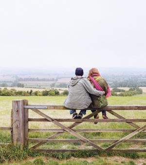 Two people sit on a fence overlooking a field. One person has an arm around the other person. 