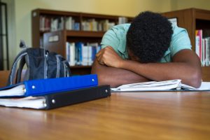 Teen hides face in crossed arms on table, binders and books open nearby