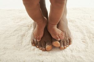 Baby feet rest on top of adult feet in sand. Both baby and adult are people of color.