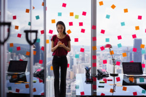 Office worker stands by glass wall covered with sticky notes writing something on list