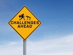 A yellow diamond sign in front of a blue sky. The sign reads "Challenges ahead" and has a silhouette leaping over a hurdle