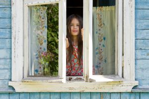 A young child with long hair pushes open window of rundown blue house