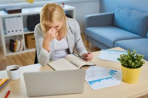 Rubbing eyes while sitting at desk in living room with documents and laptop