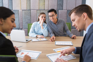 Four coworkers sit at table during meeting. Two whisper to each other