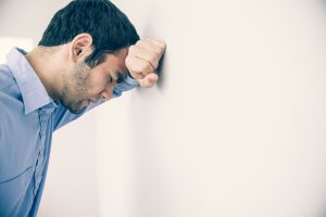 A dark-haired person in button-down shirt leans head against wall