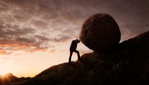 Person in silhouette pushes boulder up hill