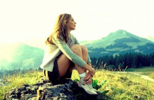 Person with long hair wearing jacket and shorts sits alone on mountaintop looking into distance