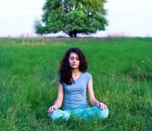 Person with long curly dark hair sits in grassy field with eyes closed, meditating peacefully