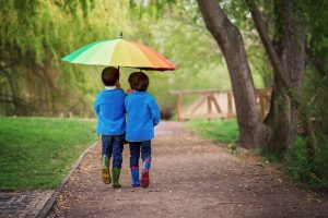 Two boys in blue jackets and rain boots walk together under rainbow umbrella