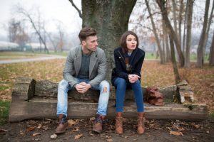 Couple sits apart on bench under tree on cold day